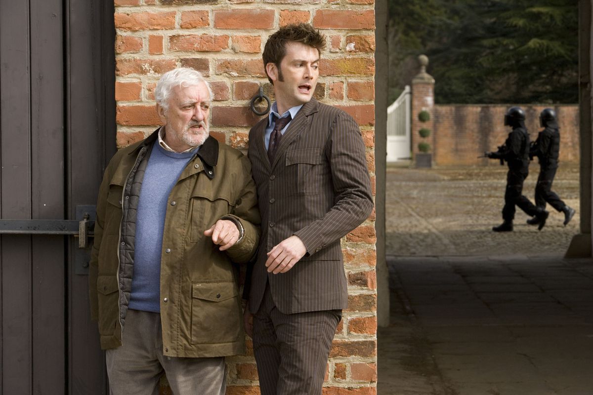 Wilfred Mott and the Tenth Doctor standing against a wall looking over Ten’s shoulder at people in black helmets walking down the alley behind them