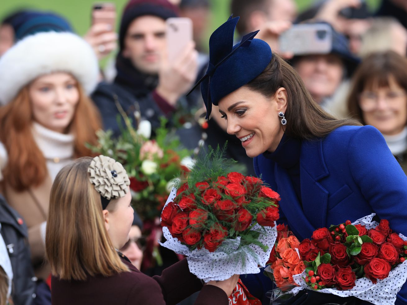 Princess Kate, in a royal blue coat and hat, bends down to accept a bouquet of red roses from a little girl amid a crowd of on-lookers.