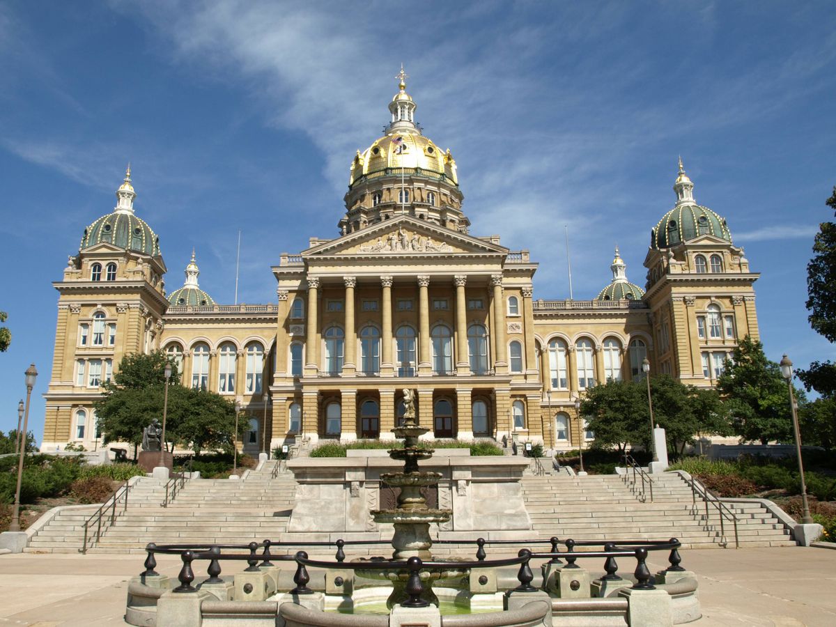 The exterior of the Iowa State Capitol Building. The facade is brown. There are multiple domed towers. The tower above the entryway is gold. There are columns in the front of the building. There is a fountain in the foreground. 