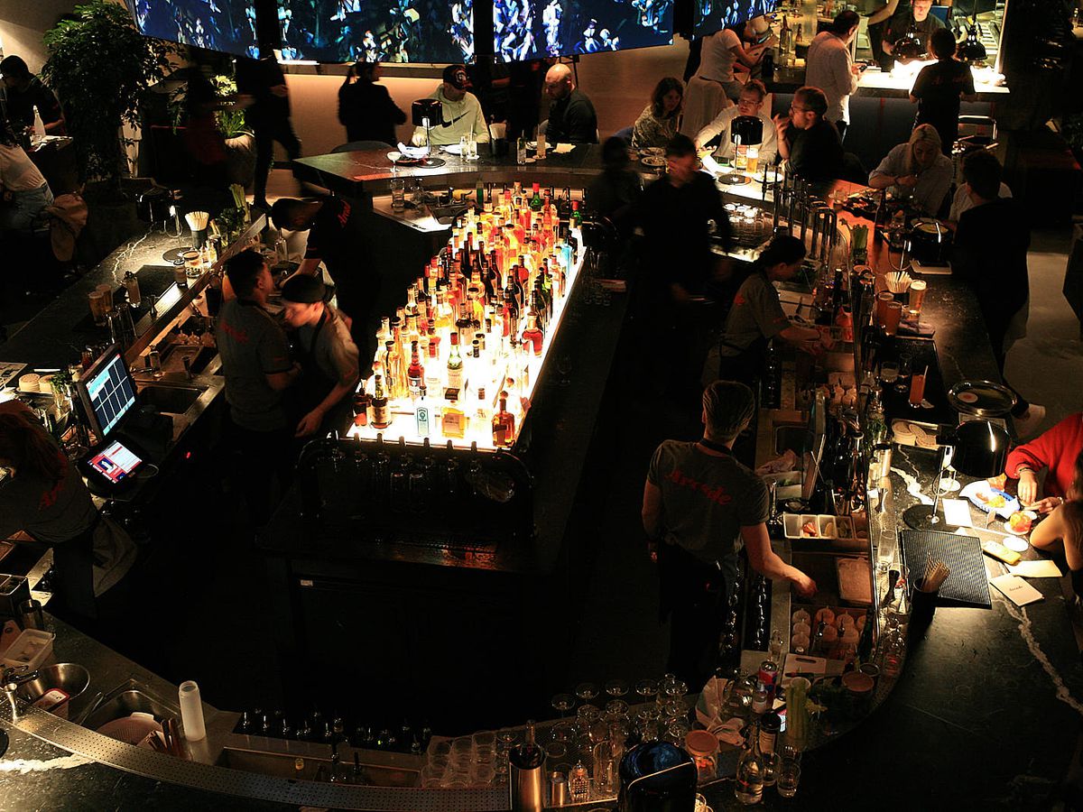 An illuminated central bar laden with bottles of alcohol, framed by large groups and booths and tables.