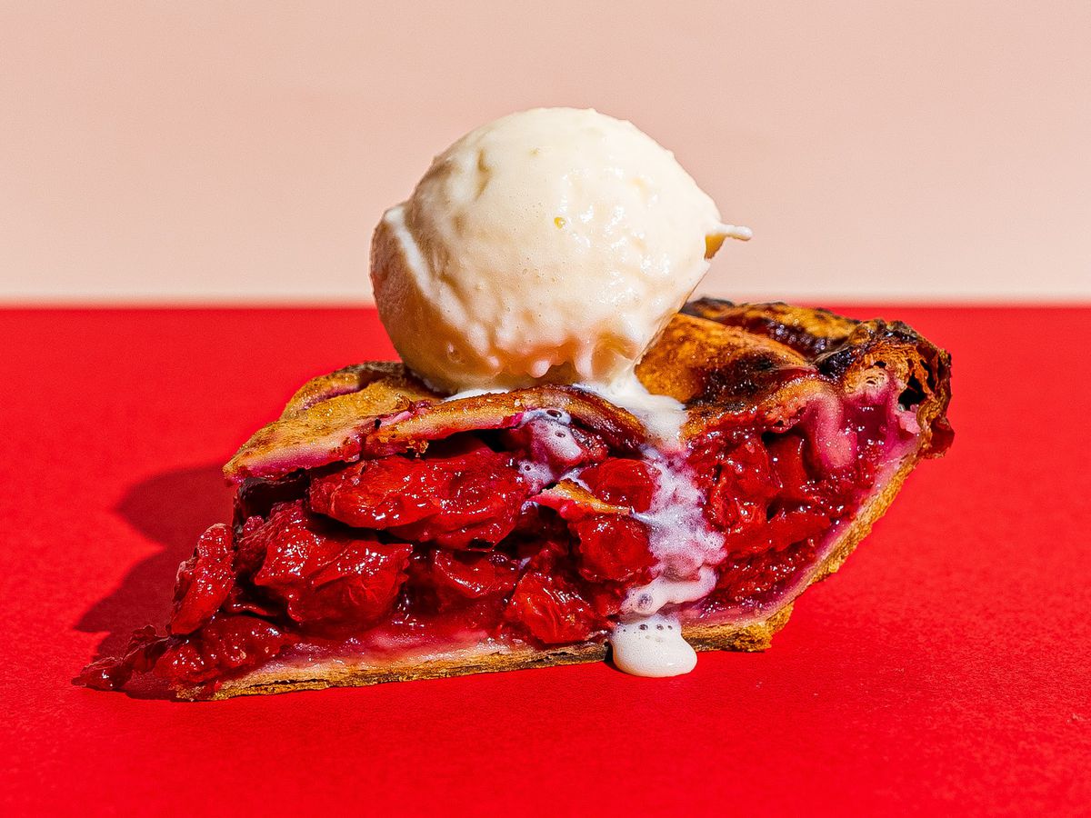 A slice of cherry pie topped with a scoop of vanilla ice cream on a red background. 