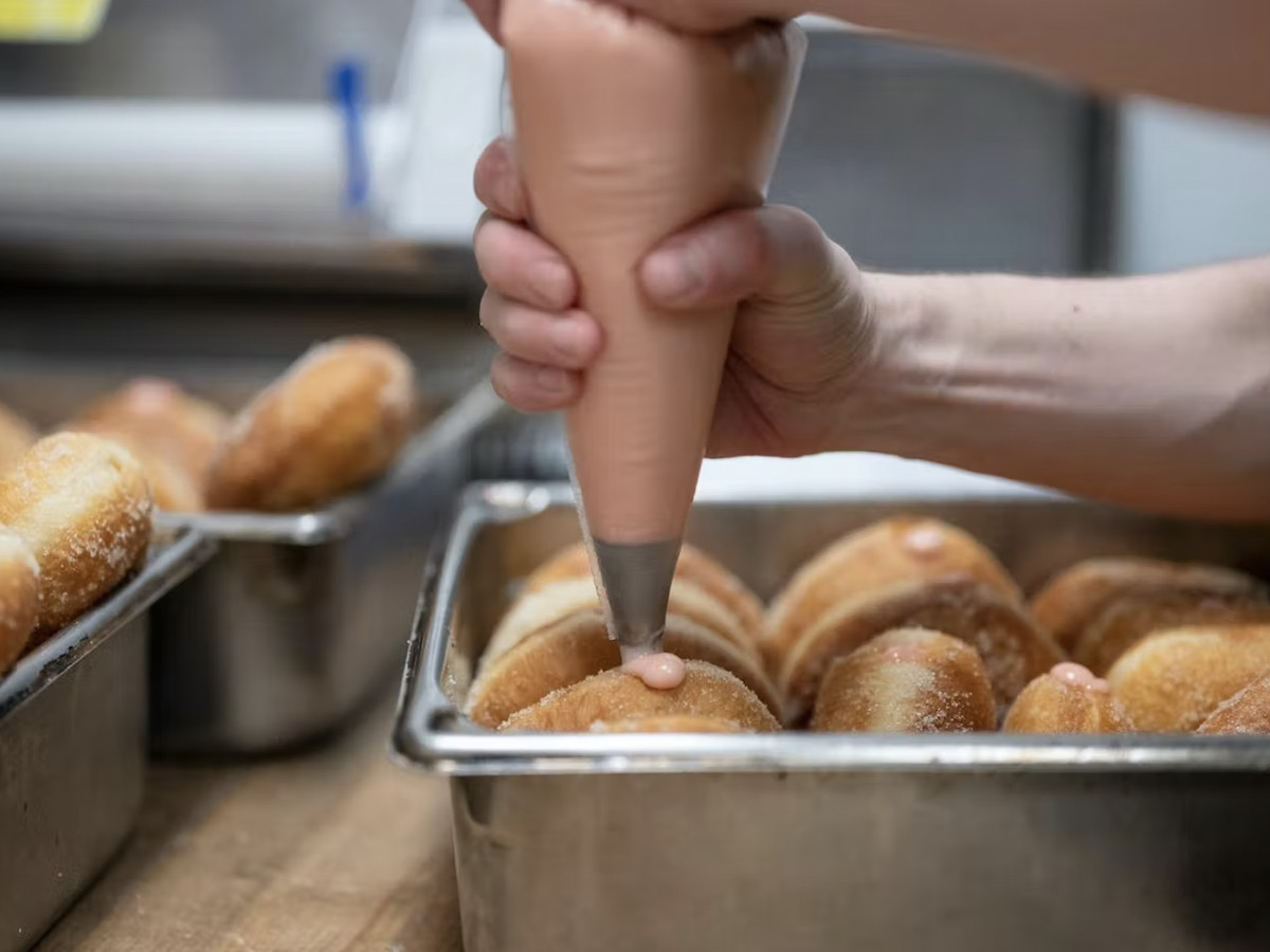 Hands hold a piping back to push icing inside rows of paczki in a metal tin.