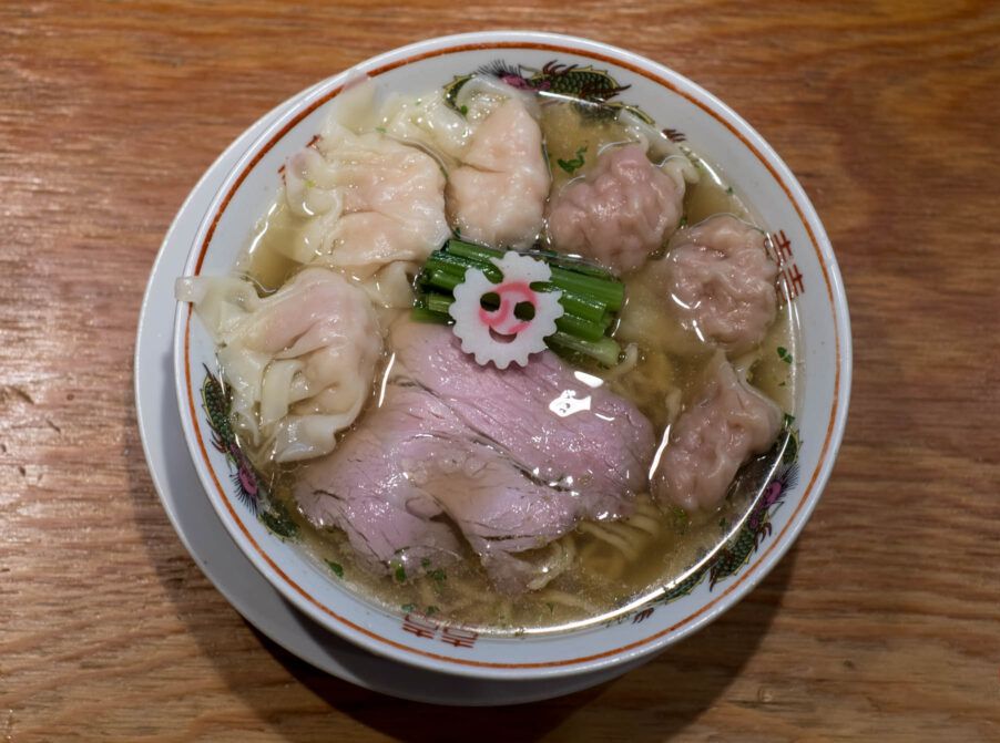 A bowl of ramen on a wood counter. The bowl includes pale slices of pork, and wontons in two varieties. 
