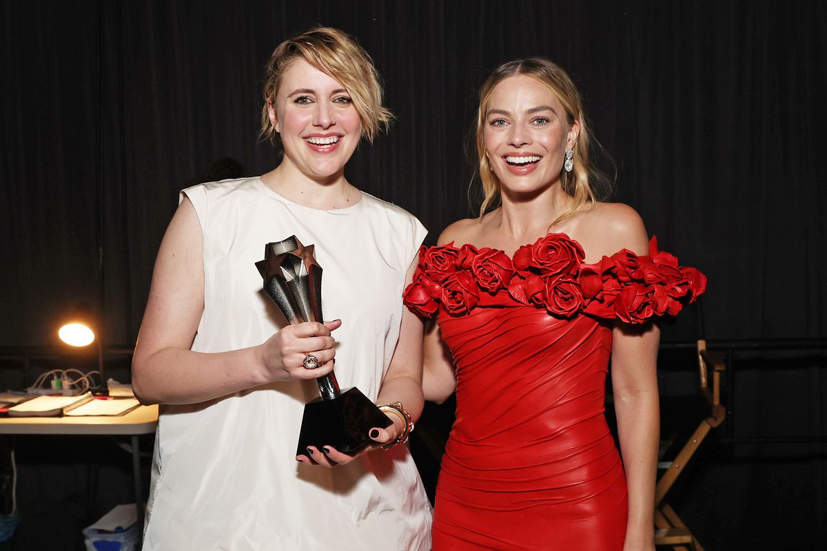 Greta Gerwig is on the left in a sleeveless white dress, with Margot Robbie on the right in an off-the-shoulder red gown. They are both smiling and holding an award.