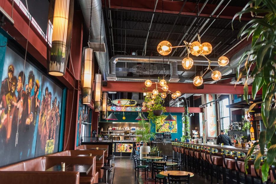 A light-filled restaurant interior with industrial style chandeliers with exposed bulbs, a bar with leather swivel seats, leather booths, a mural on one wall and colorful decorations throughout