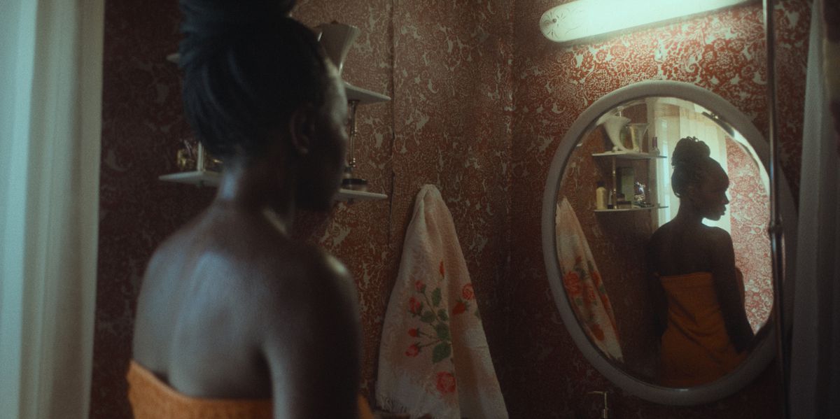 Aisha (Anna Diop), a dark-skinned woman wearing a bright orange towel, examines herself in a mirror in a darkened room in Nanny