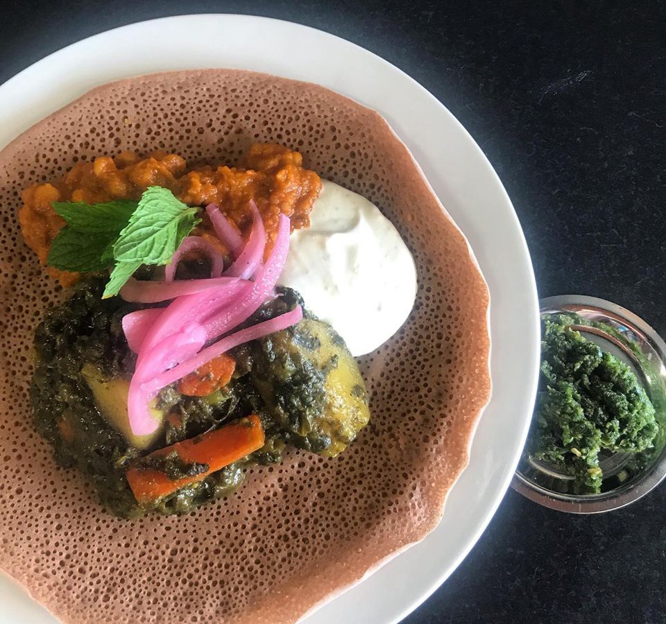 As seen from above a plate nearly filled with spongy injera flatbread, topped with a colorful curries and yogurt beside a small bowl of green dip