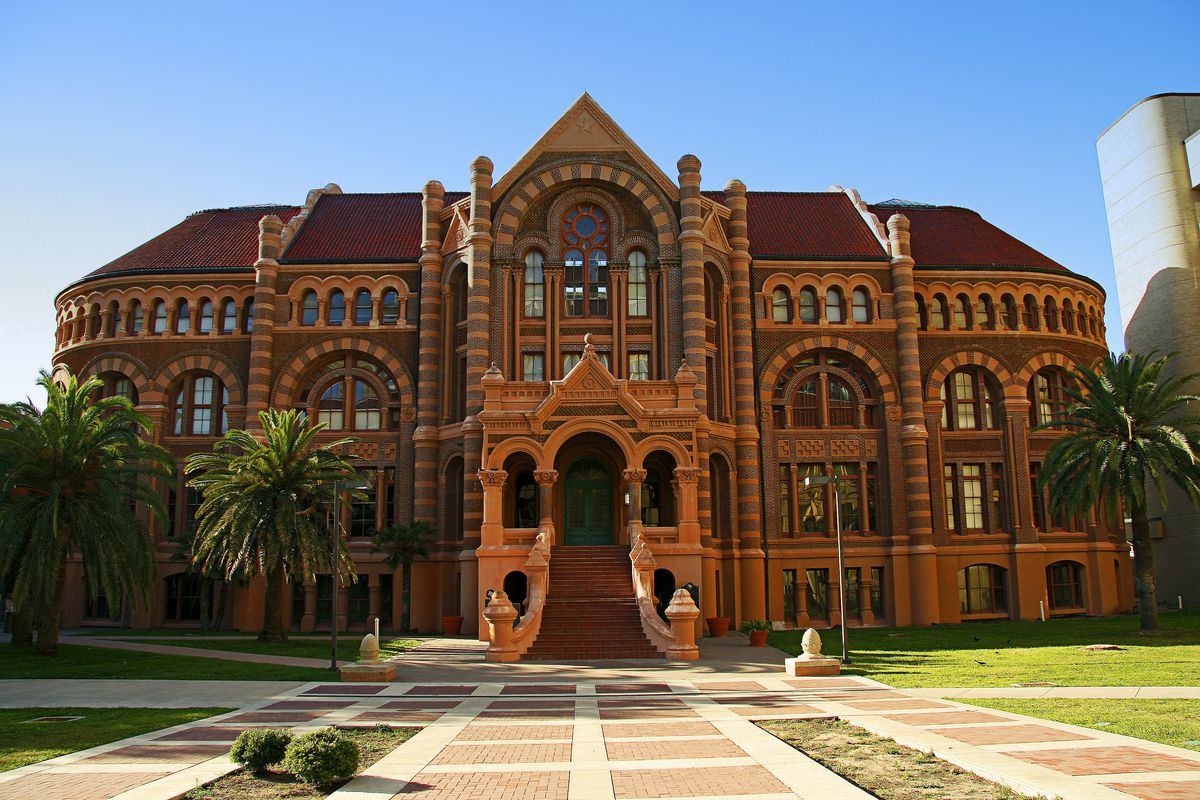 The Ahbel Smith Building at the University of Texas. The building’s facade is orange and red stone with a staircase leading up to the door. There is an arched structure over the door.