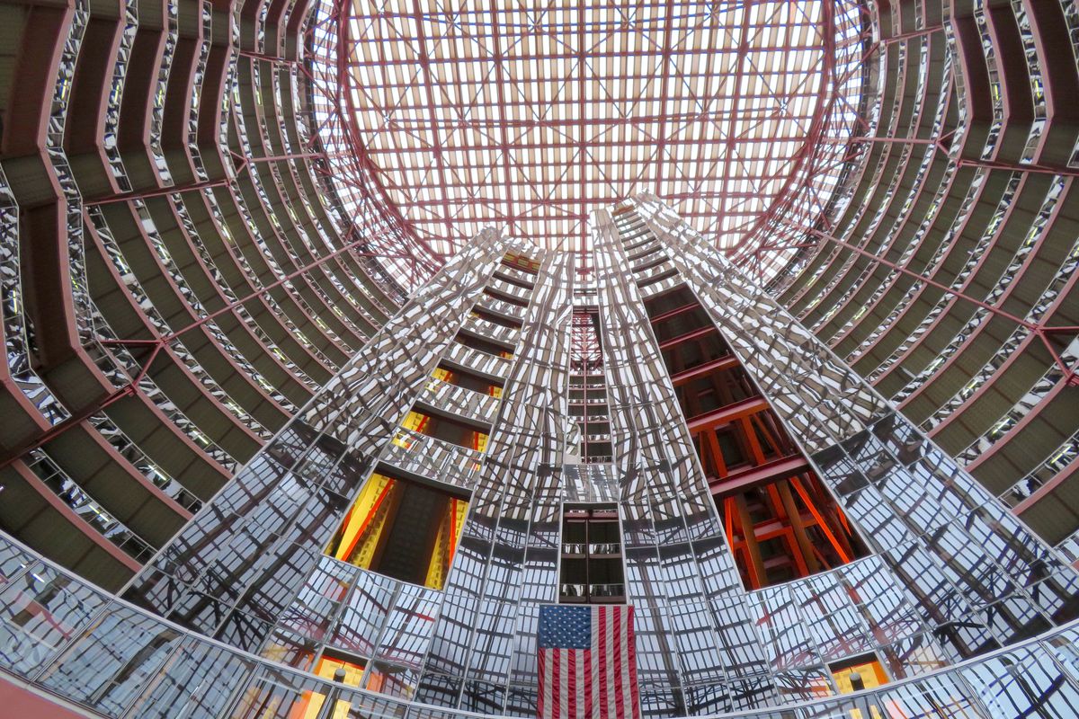 A view upward at a massive circular Thompson Center atrium ringed with offices. Glass elevator shafts rise in the center and reflect the surrounding architecture.