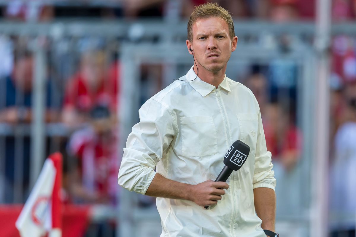 Julian Nagelsmann holding the DAZN mic on the sidelines as he gives an interview for the match between Bayern and Wolfsburg. He looks slightly annoyed.