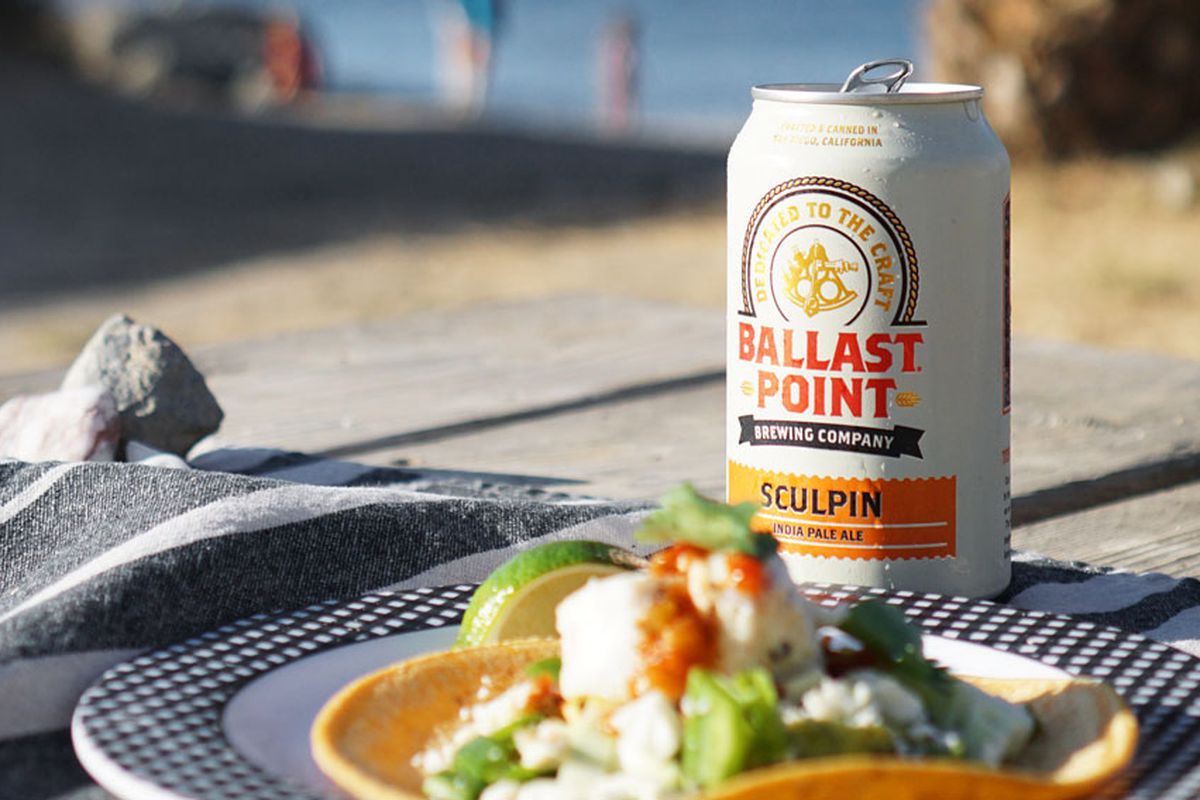 A can of Ballast Point beer on a wooden table beside a plate of food.