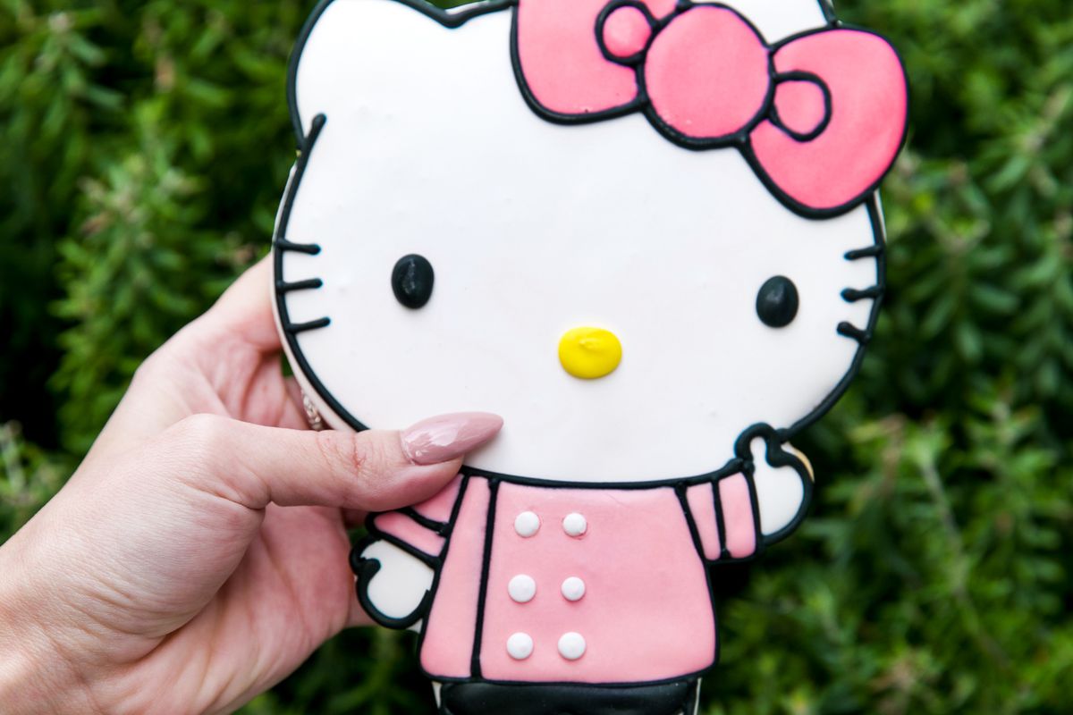 A white woman’s hand with long pink nails holds an oversized Hello Kitty cookie. Hello Kitty wears a pink smock top and a matching pink bow, with a yellow nose, white face and black whiskers.