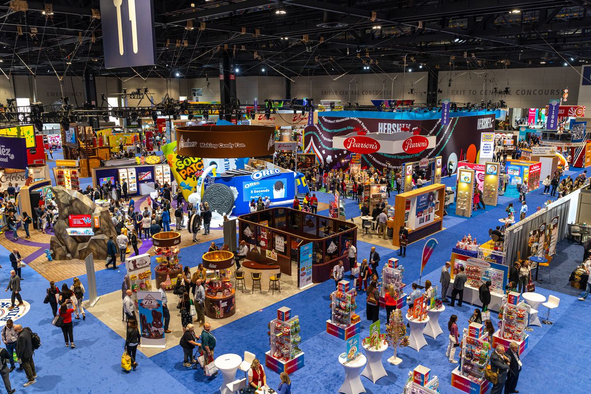 A crowded convention floor with blue carpeting and snack food companies’ promotional stalls scattered throughout.