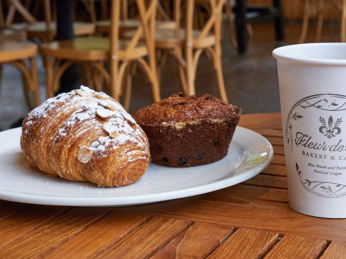 An almond-topped croissant and a muffin on a plate next to a paper cup full of black coffee.
