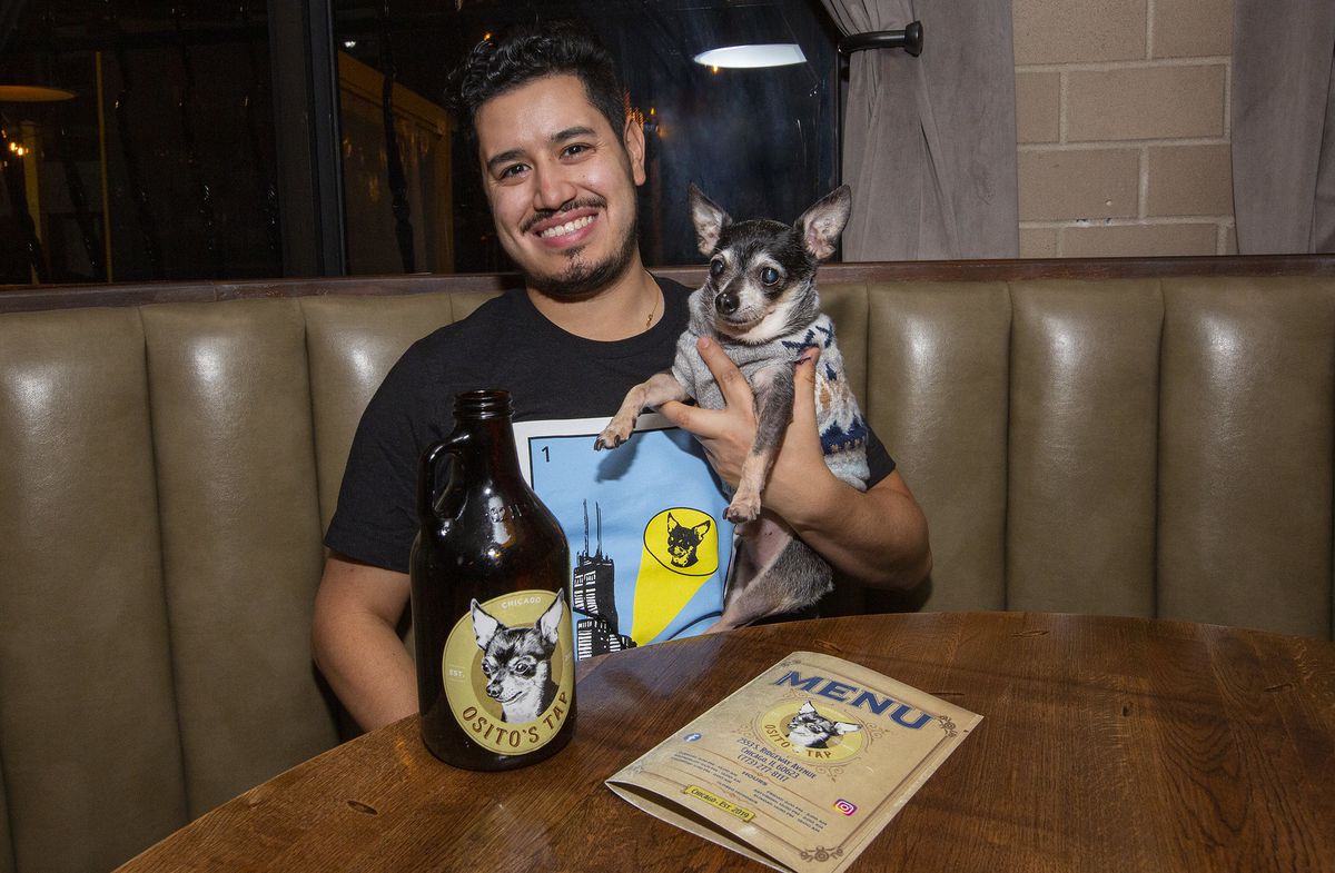 A smiling man and a chihuahua in a sweater sit together in a brown vinyl booth with a growler of beer and a menu