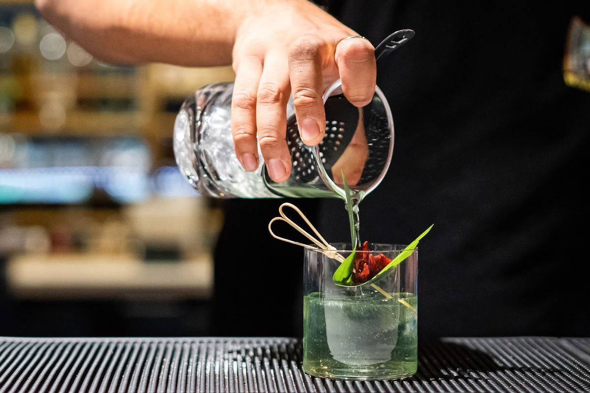Bartender pouring a clarified pandan cocktail into a short circular glass that holds a cylindrical ice cube and flower.