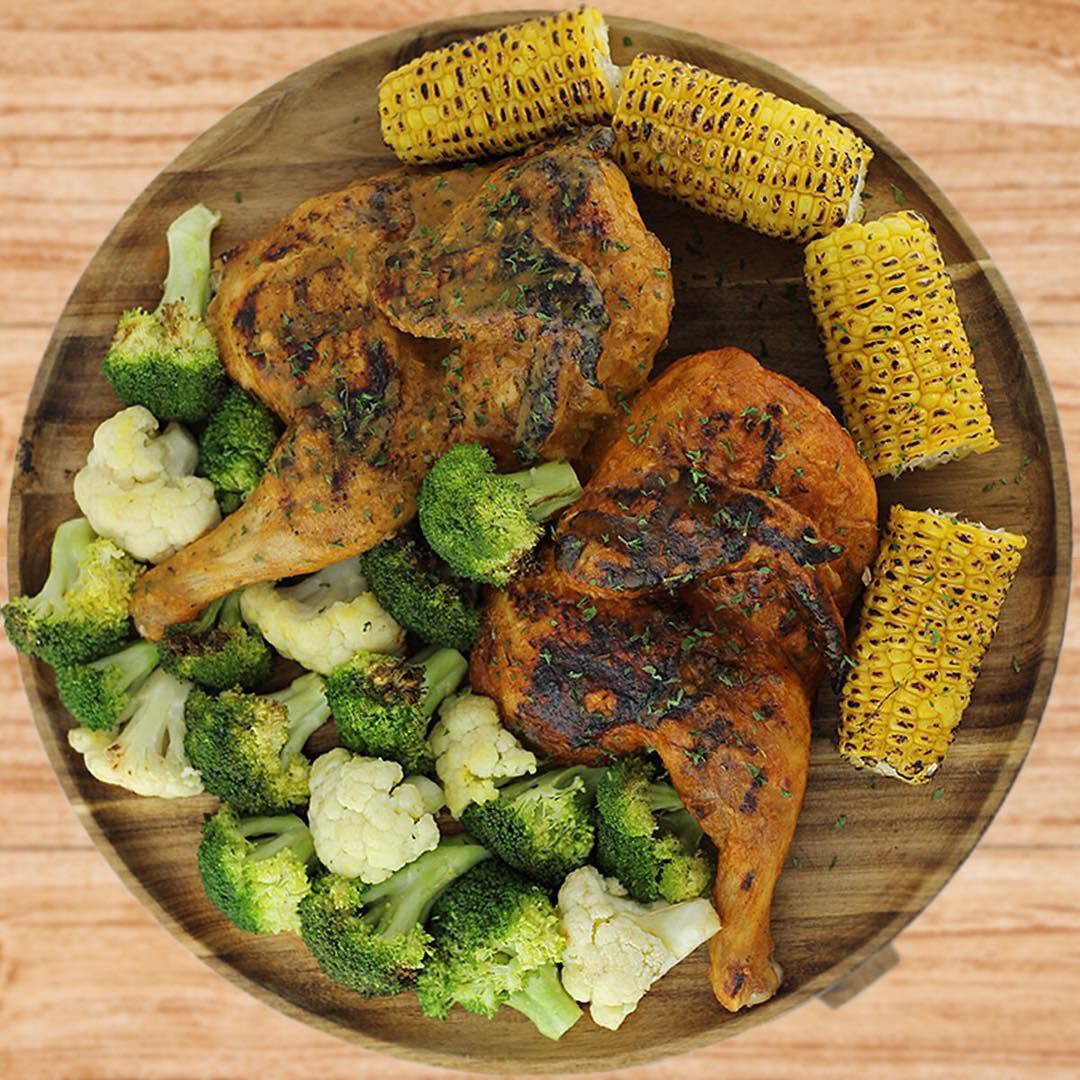 A wooden plate with halved roasted chicken, broccoli and cauliflower, and pieces of corn.