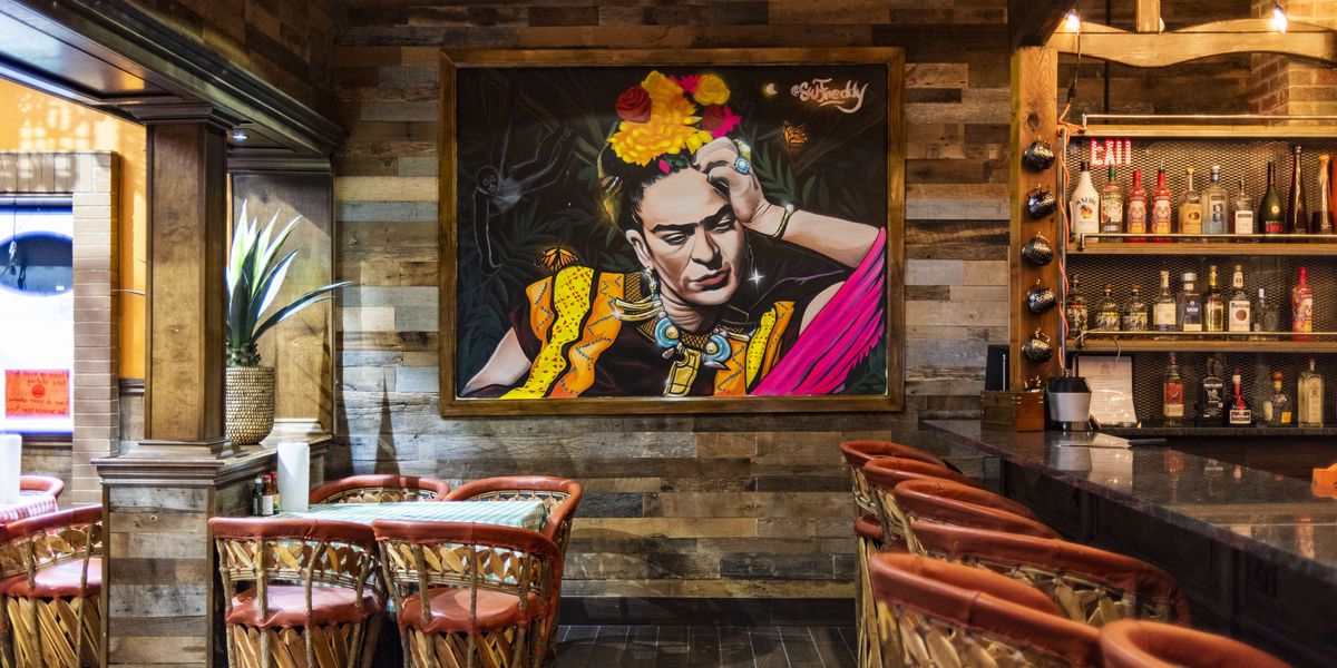 A painting of Frida Kahlo on a wood paneled wall with several chairs and a bar.