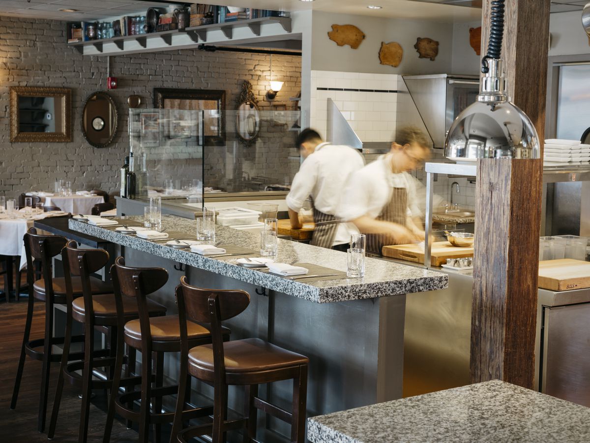 Two cooks work in the open kitchen at Craigie on Main in Cambridge, with a marble bar and high-top chairs in front of it and part of the dining room visible in the background.