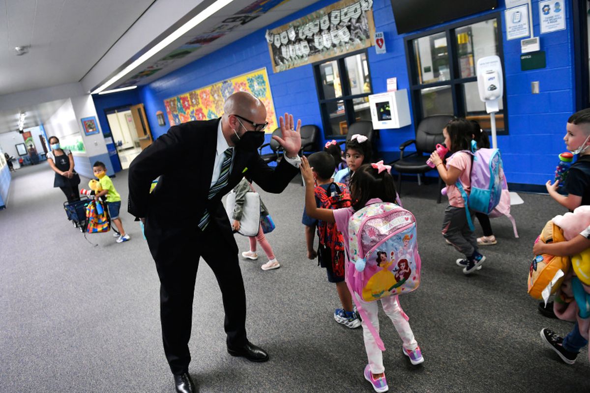Denver schools Superintendent Alex Marrero high-fives a young student wearing a pink backpack in a school hallway. The superintendent is wearing a suit and a face mask.