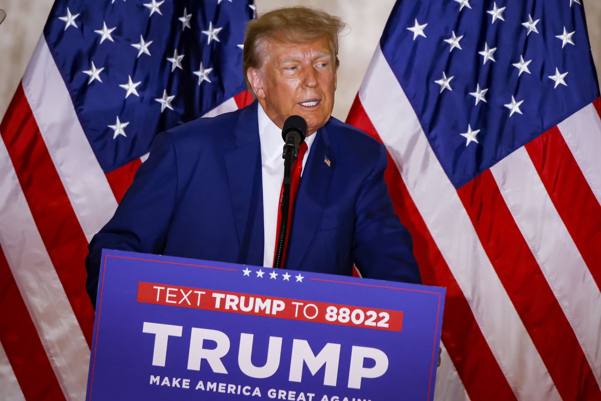 Donald Trump speaks at a wooden podium with a “Trump” campaign sign on it, two large American flags hanging behind him.