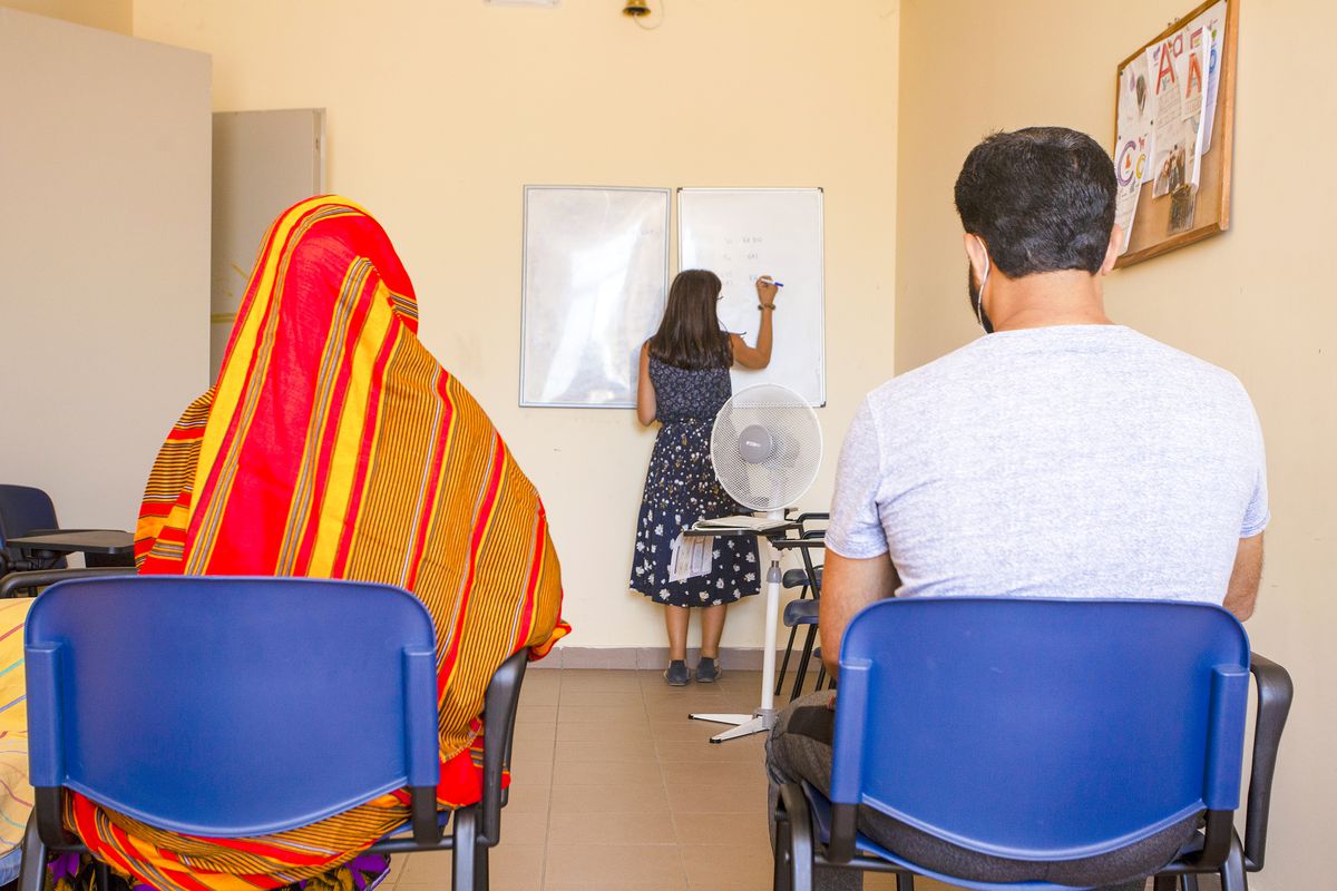 Two adult students sit in a classroom and watch an instructor write on a whiteboard.