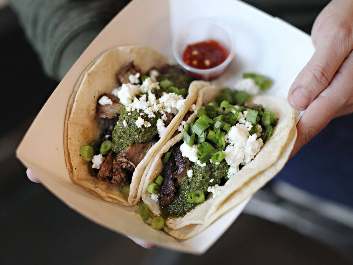 A hand holds a paper boat of two tacos topped with green sauce