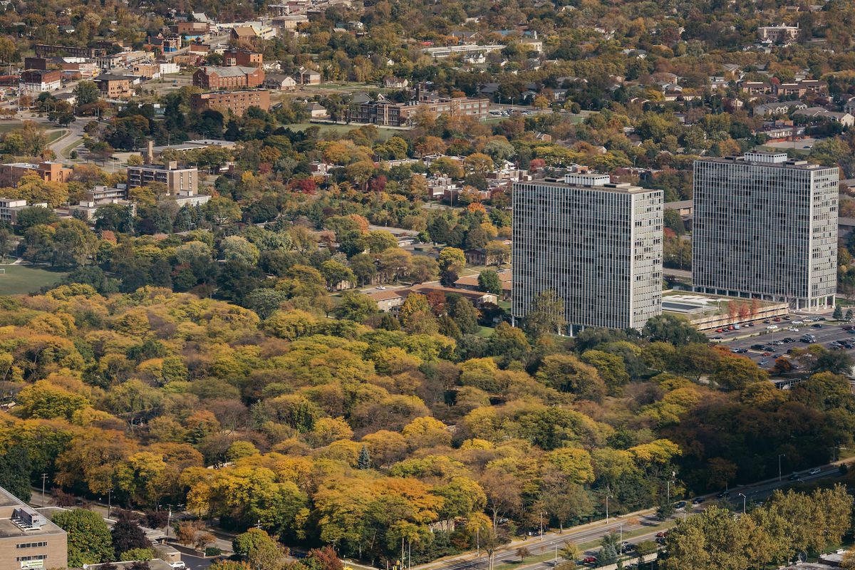 An aerial view of a large, tree-filled park. Two metal towers rise on the right side.
