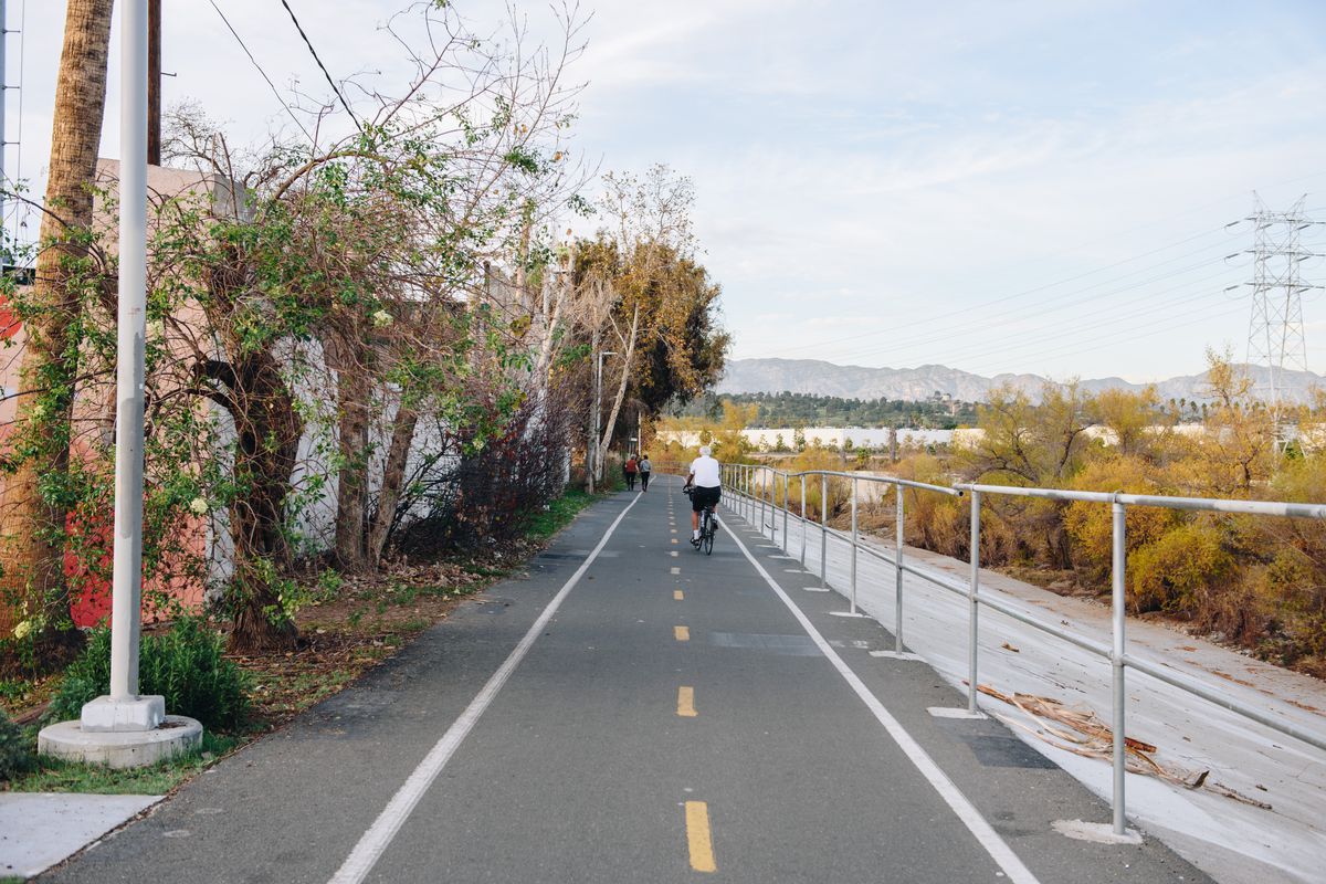 A cyclist rides along an open bike path that abuts a mostly dry river.