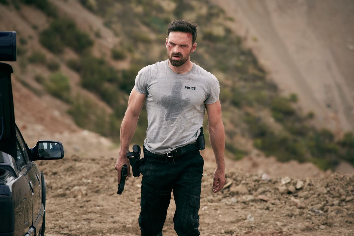 A man in a sweat-soaked t-shirt and black cargo jeans with bloody scars on his face holds a pistol and walks beside a black car in a rocky desert.