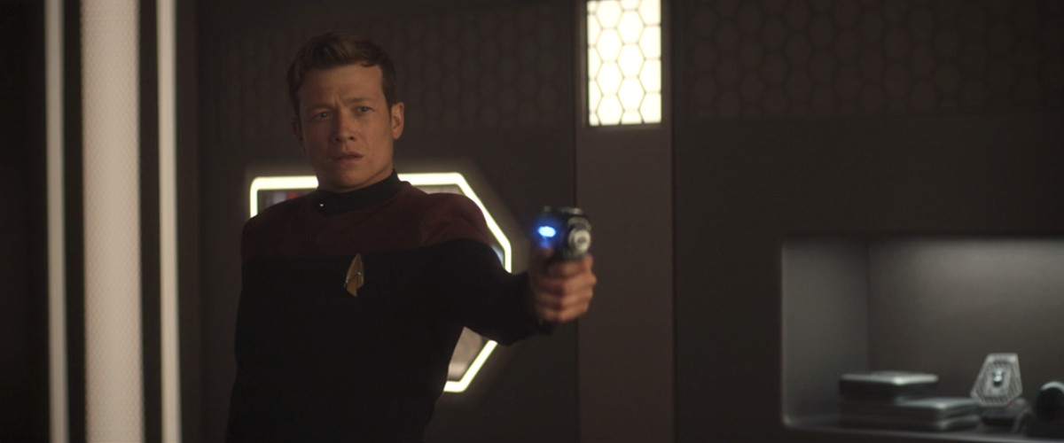 Jack Crusher, in his Starfleet ensign uniform, standing in his quarters, raises his phaser and looks confused in Picard. 