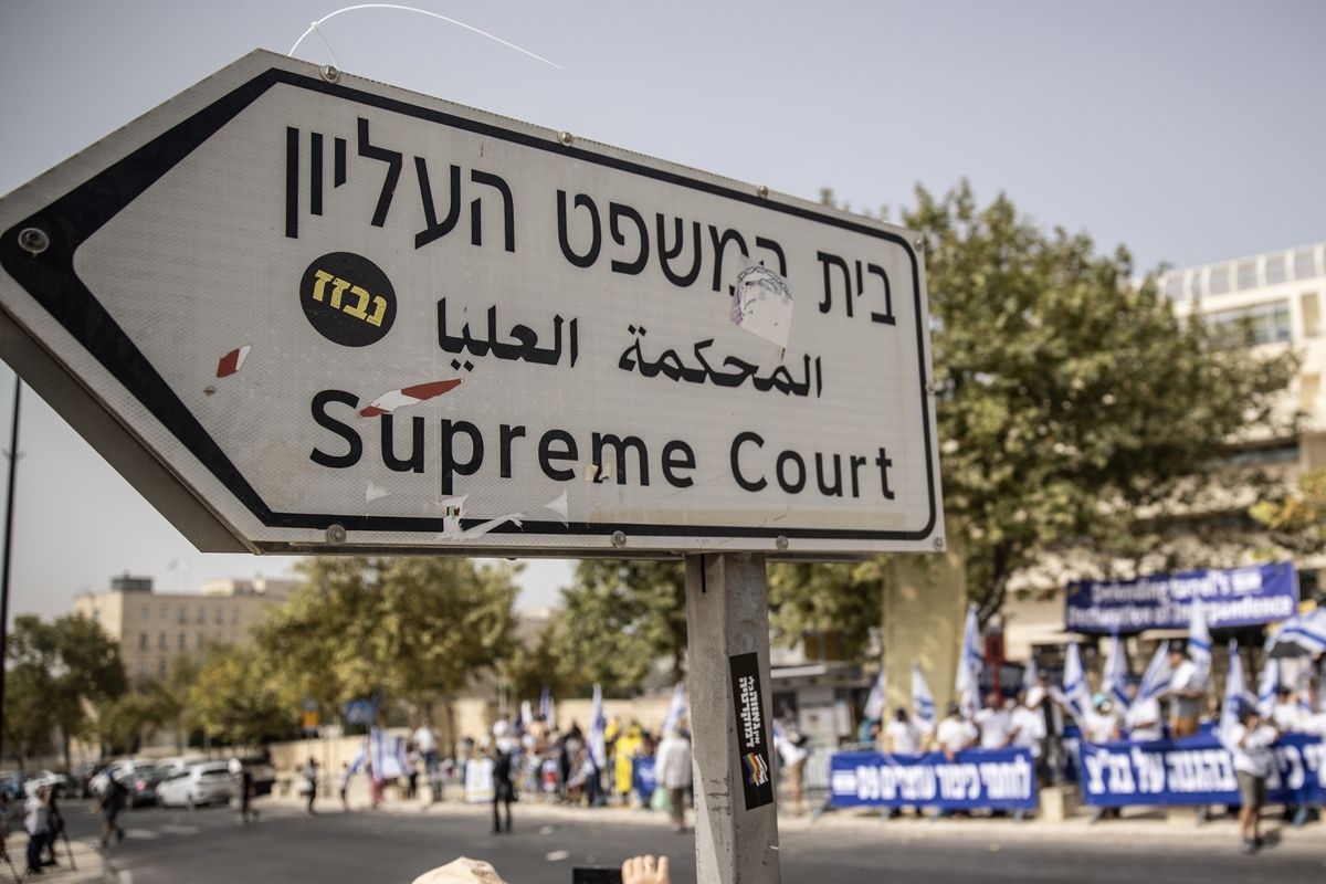 A sign pointing to the Supreme Court in Jerusalem is backdropped by dozens of people on the nearby sidewalks with banners and signs.