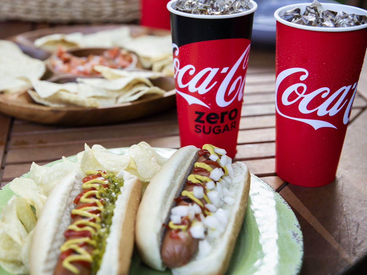 Two hotdogs on a plate, with two paper cups of Coca-Cola beverages. 