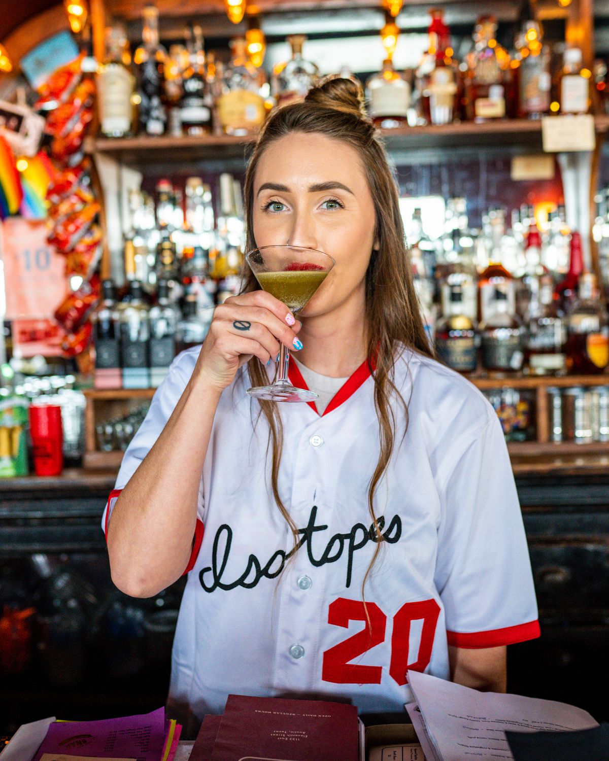 A woman in a white baseball jersey with red trim that reads Isotopes 20 sipping a dark green cocktail in a martini glass.