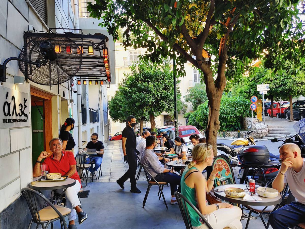 Sidewalk seating outside a restaurant, with patrons seated at small tables and servers passing through