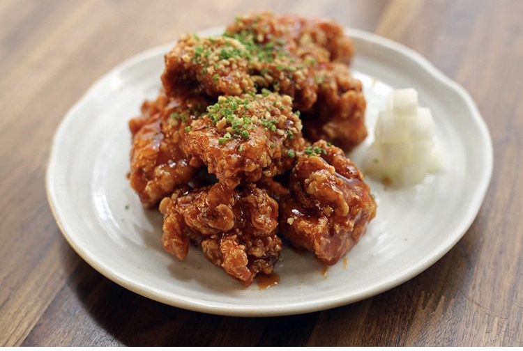 A pile of reddish brown fried chicken with green garnishes sits on a white plate with a small pile of cubed, white pickled daikon radish sitting next to the chicken. The plate is set on a light wooden table.