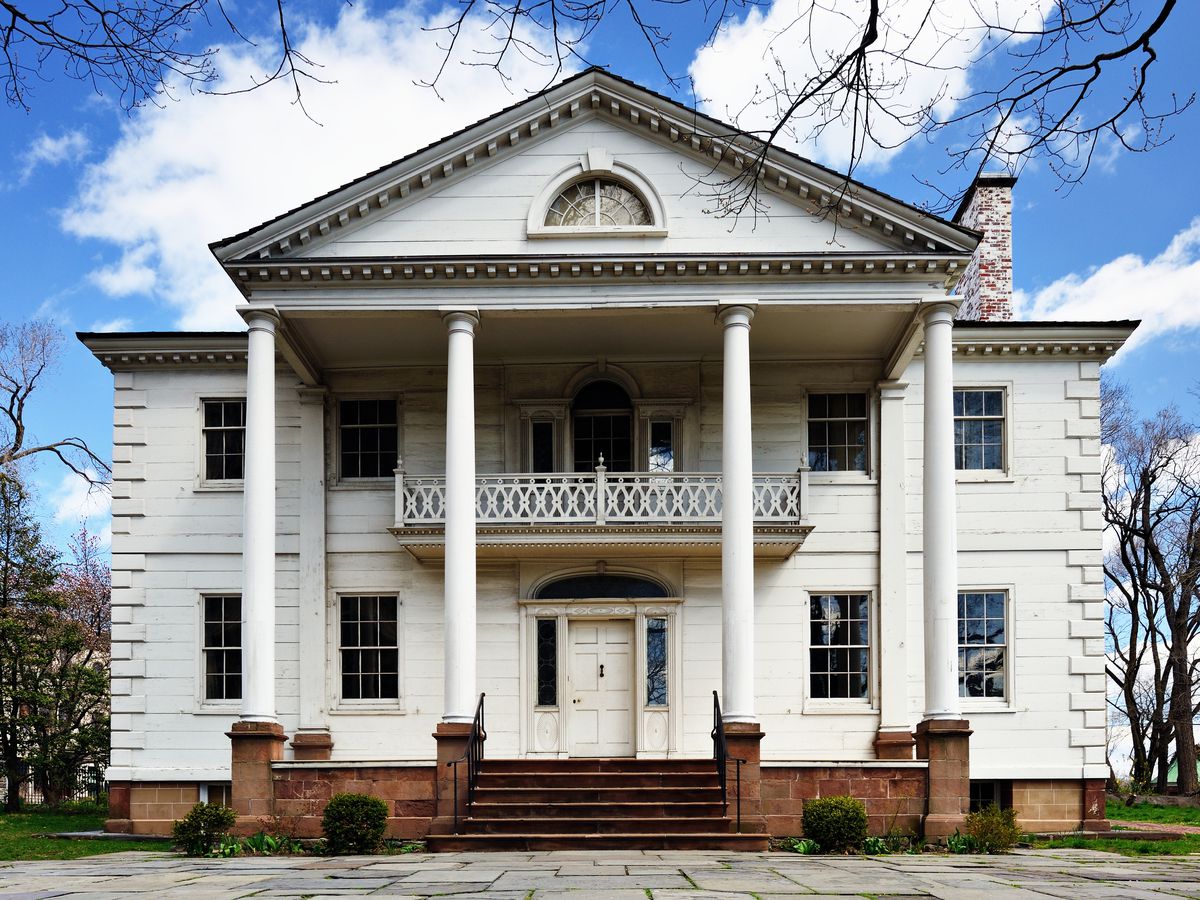 The exterior of the Morris Jumel Mansion in New York City. The facade is white and there are columns and a balcony on the front of the building.