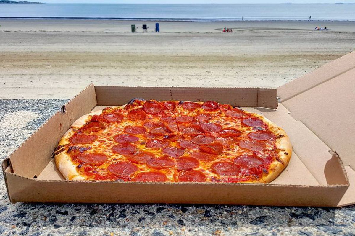 A pepperoni pizza sits in an open cardboard pizza box on a stone surface, with a mostly empty beach visible in the background