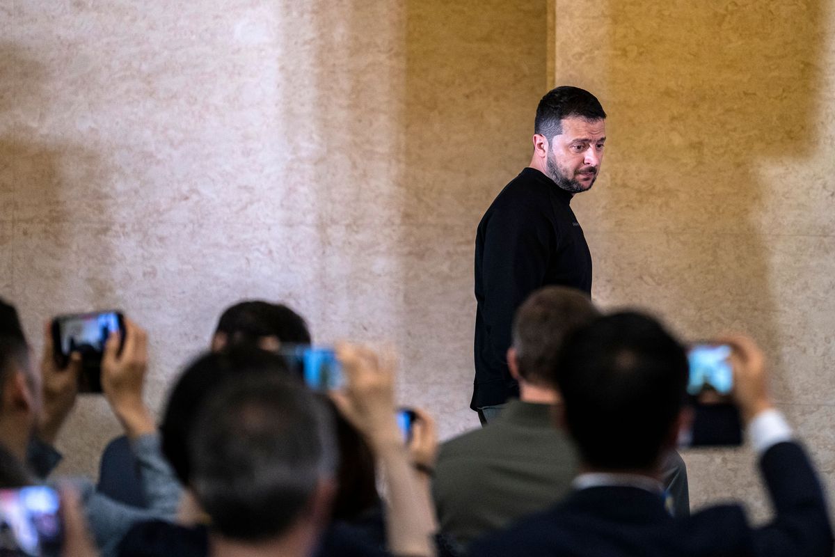 Zelenskyy, wearing a black turtleneck, walks past a group of seated people filming him with their Iphones.