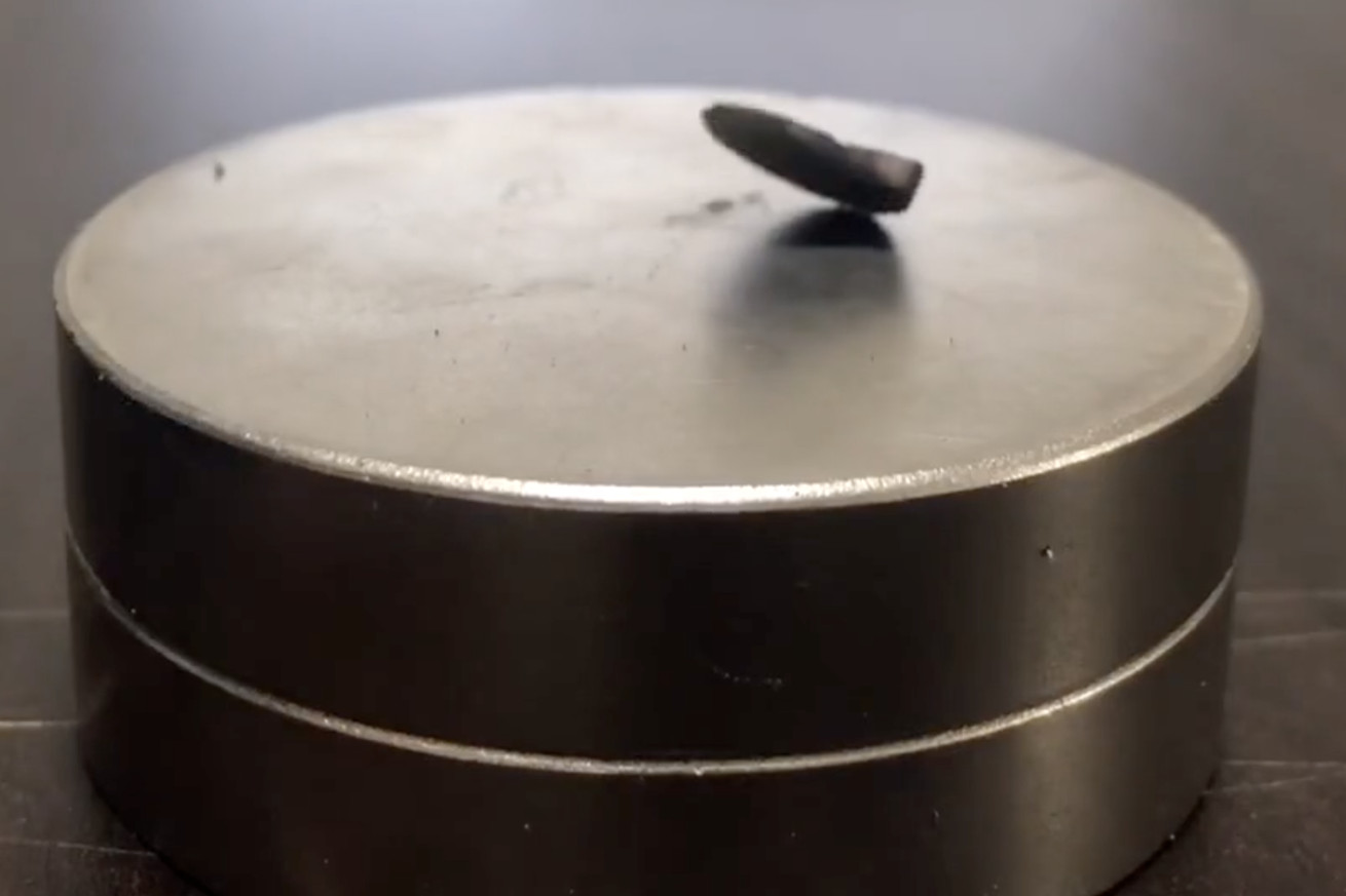 A screen shot of a fragment of dark gray rocky material partially floating over a round magnet. One side of the fragment is raised in the air while the other is touching the magnet.