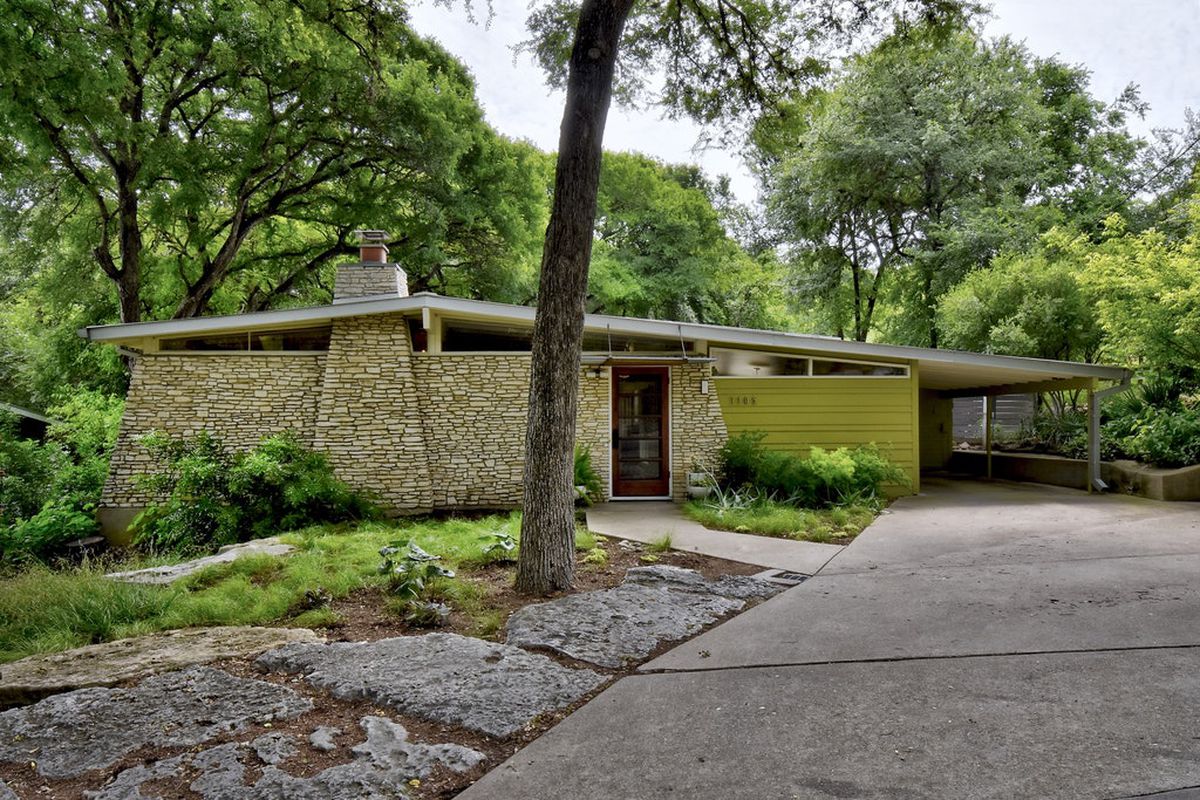 An exterior view of a midcentury modern house in Austin. There is a concrete driveway, a stone wall, and a green exterior wall. 