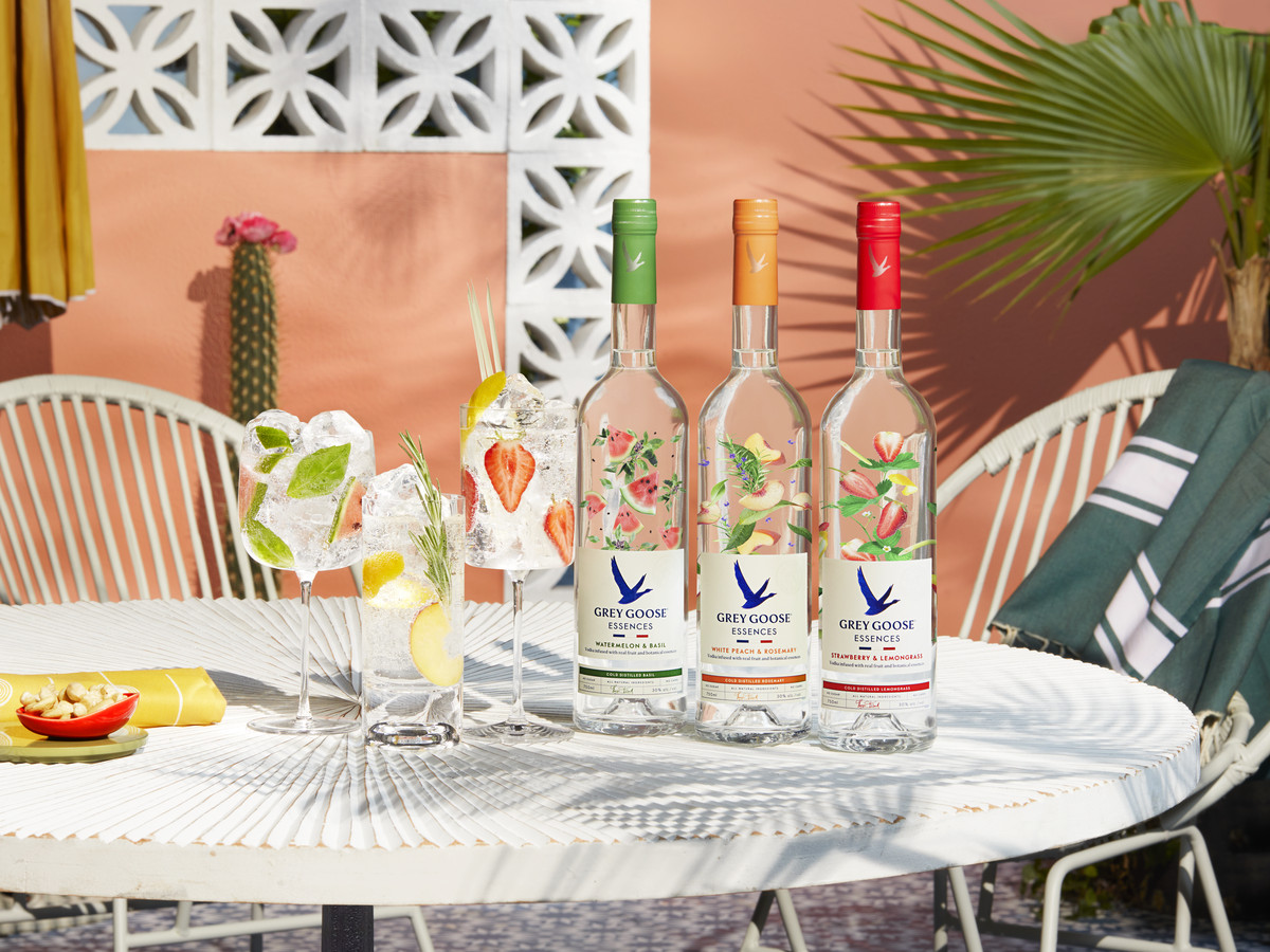The three Grey Goose Essences flavors on a white patio table outdoors with a palm frond and cactus in the background.