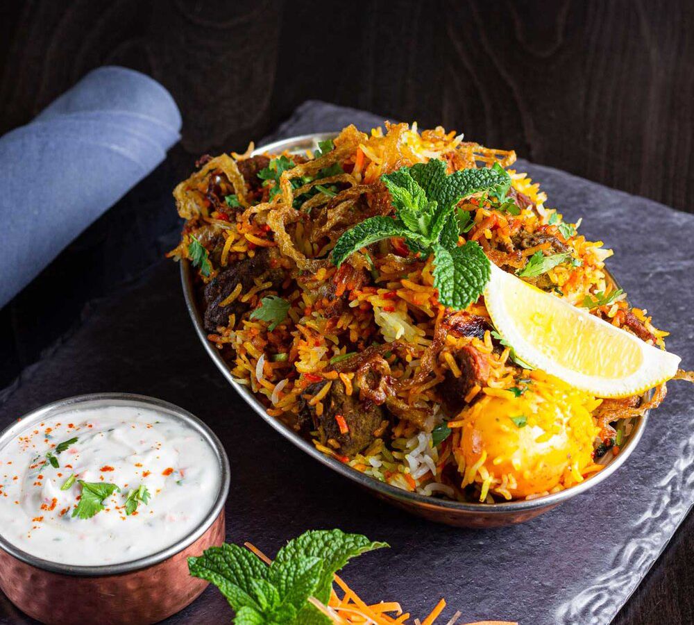 A metal dish overflowing with biryani, studded with chunks of dark meat, topped with mint and a lemon wedge. Served on a slate board with a small tin raita on the side
