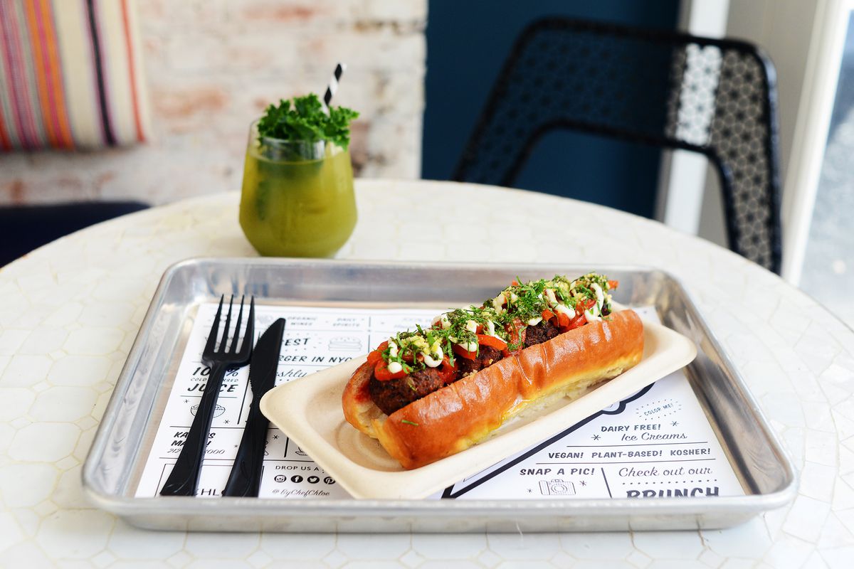 Meatballs sit in a glossy hot dog-style roll on a metal tray that’s covered with a white placemat with a lot of text on it in a modern font. There’s plasticware on the tray, which sits on a white table.
