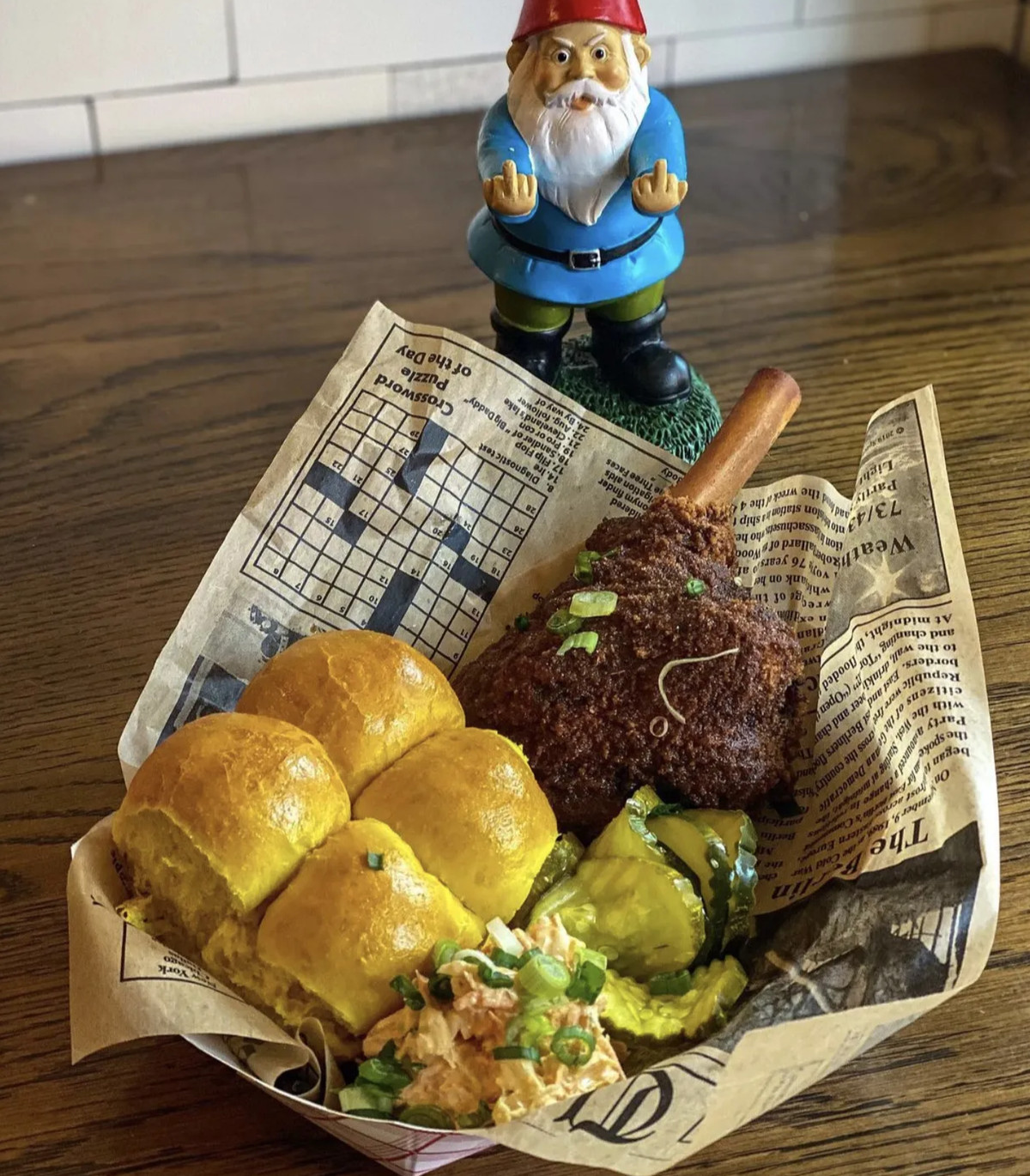 A basket of Nashville hot lamb shank served with Hawaiian rolls, pickles, and slaw with a gnome statue in the back