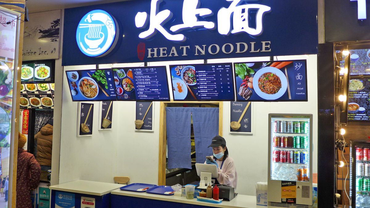 The exterior of a food stall with blue signage. A woman can be seen working at the cash register behind a counter