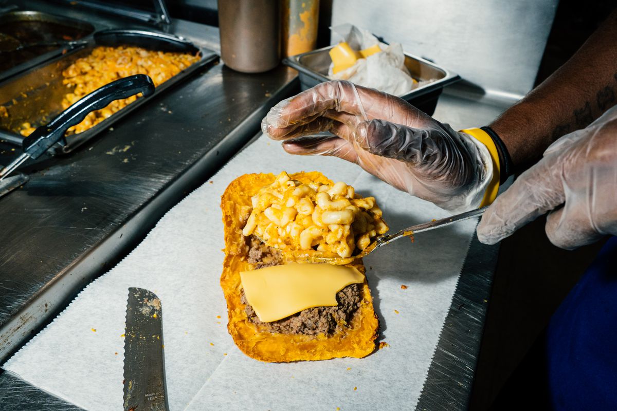 A gloved hands scoops baked mac and cheese onto a beef patty with American cheese.