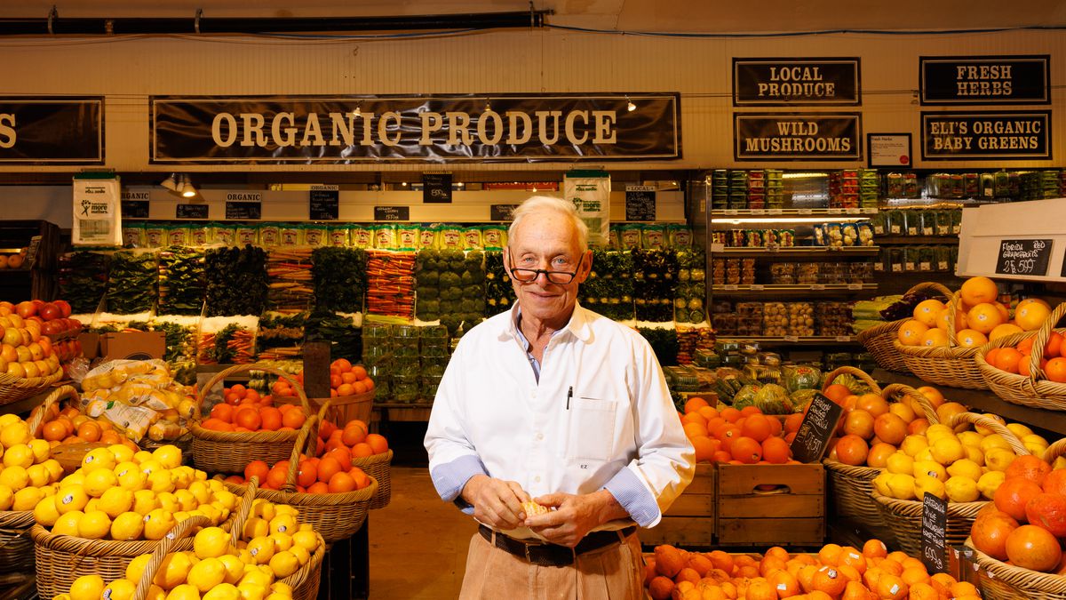A man in a white shirt and khakis stands among produce at his restaurant. 