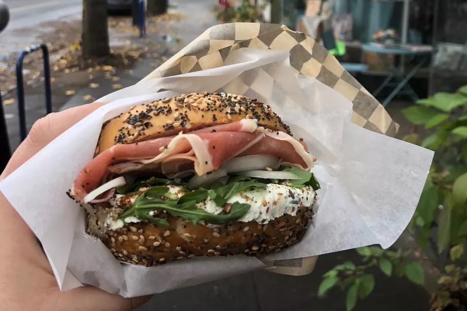 A picture of a hand holding a bagel with prosciutto, arugula, onions, and cream cheese