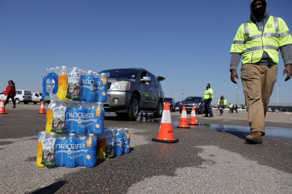 A Black man in a reflective neon vest walks toward piled cases of water, as cars line up behind him.